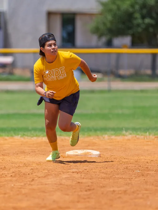 A player wearing a yellow "Pride Sports USA" shirt, dark shorts, neon socks, and a backwards cap is running on a kickball field, appearing to round a base during gameplay.