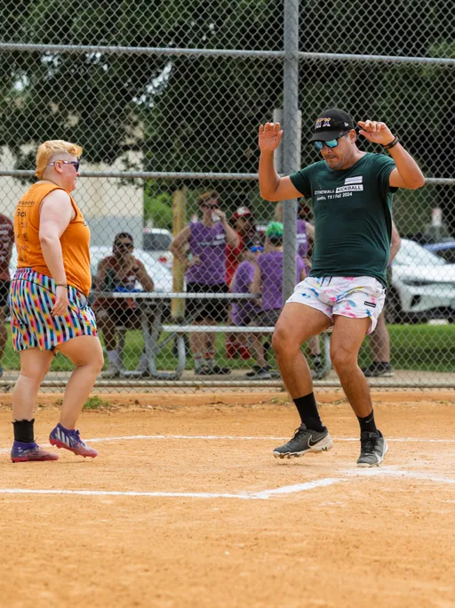 Two kickball players are at a base on a dirt field; one player is wearing an orange shirt and rainbow shorts, and the other is in a green shirt, patterned shorts, a hat, and sunglasses, with arms raised in celebration. Spectators are visible behind the fence in the background.