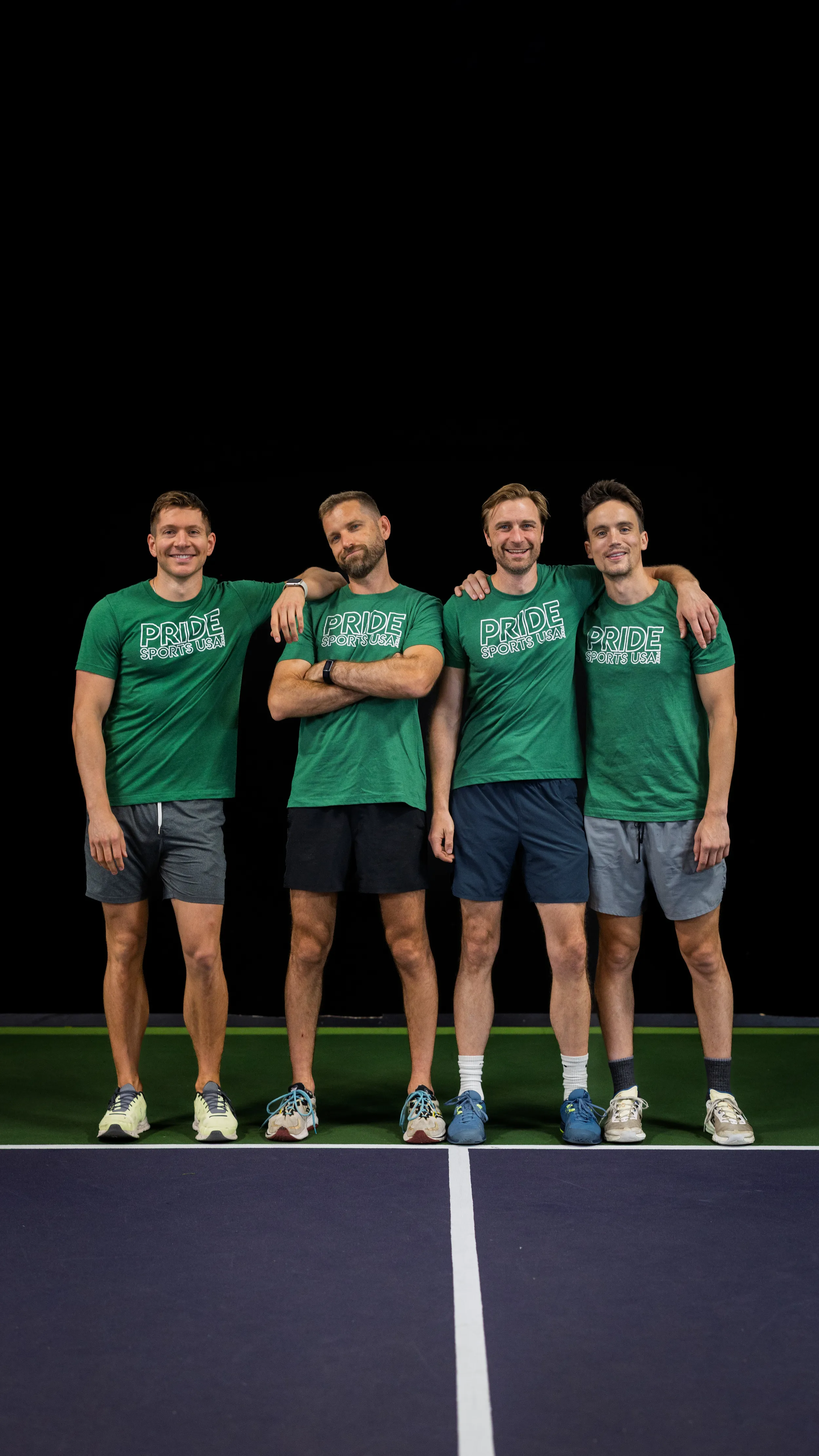 Four players stand arm-in-arm on an indoor pickleball court, posing near the center line. They wear matching green “PRIDE SPORTS USA” T-shirts with athletic shorts and sneakers against a dark backdrop.