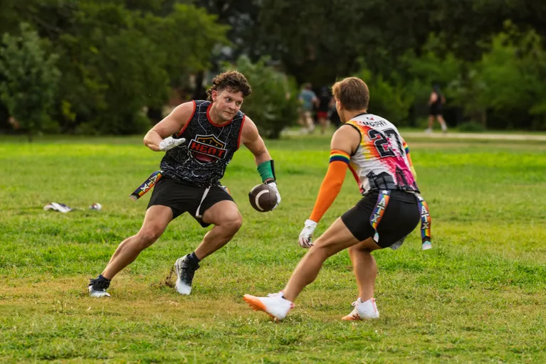 During ATX Heat (Orange) practice for Gay Bowl 2025, two flag football players scrimmage on a grassy park field—one in a black “HEAT” jersey with a green arm sleeve plants to cut with the ball while a defender in a white-and-orange top with a rainbow armband reaches to pull the flag, with trees and a few people in the background.