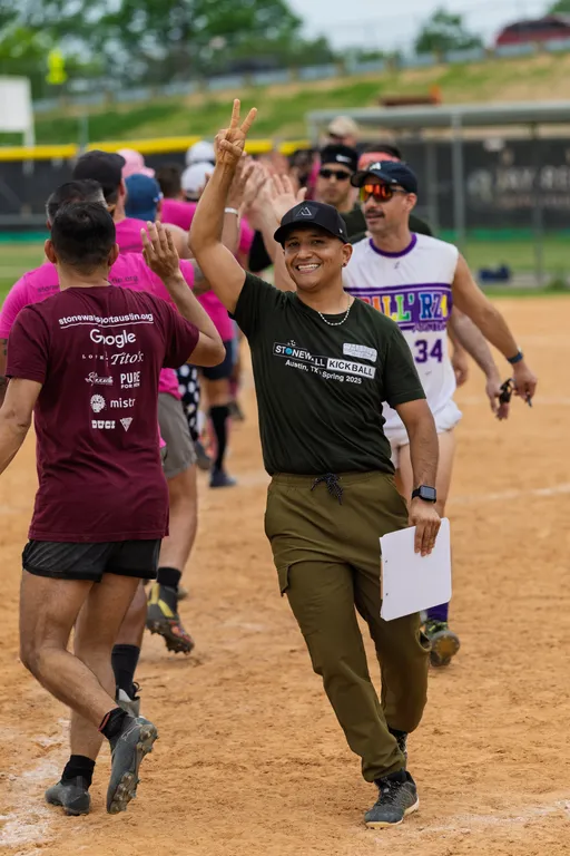 Players on a kickball field give each other high fives in a post-game line; one person in front, wearing a "Stonewall Kickball" shirt, a cap, and holding a clipboard, is smiling and making a peace sign gesture.