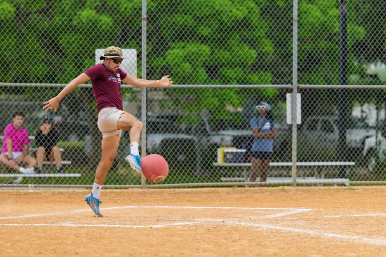 A kickball player in a maroon shirt, light shorts, and patterned hat is captured mid-kick on a sandy kickball field, while several people watch from behind a chain-link fence.