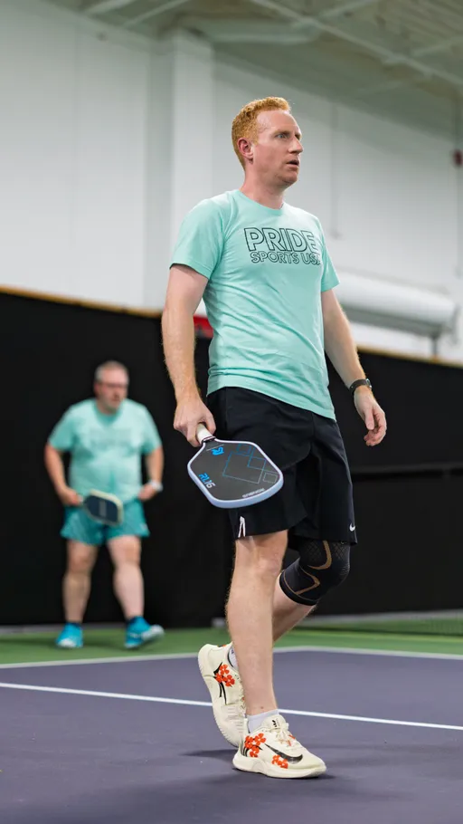 Two players are on an indoor pickleball court, both wearing mint green "Pride Sports USA" shirts and holding paddles; the player in the foreground is standing at the edge of the court with a knee brace and decorated sneakers, while another player stands ready in the background.