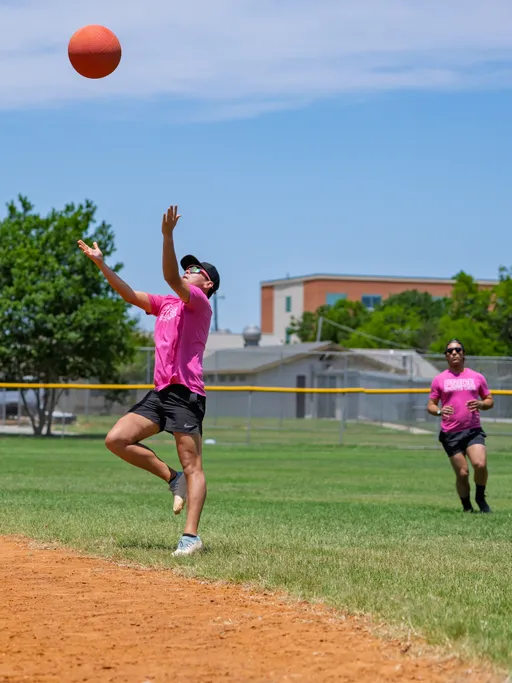 A kickball player wearing a pink shirt, black shorts, and a cap is preparing to catch a ball in mid-air, while another player in similar attire runs toward the action on a grassy field.