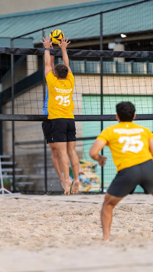 Two sand volleyball players in yellow shirts, one jumping high at the net with arms outstretched to block the ball, while another player in yellow stands nearby on the sandy court; a volleyball net, tall fence, and buildings are visible in the background.