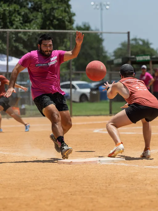 Two kickball players are in action on a dirt field, one wearing a bright pink shirt sprinting towards a base, while another in an orange shirt and black cap prepares to catch the ball; a red kickball is mid-air between them with other players and trees in the background.