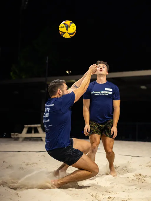 On a lit sand volleyball court at night, a player in a navy Stonewall league shirt drops to their knees and attempts a set on a yellow-and-blue ball, kicking up sand as a teammate in a matching shirt and patterned shorts waits ready behind; a picnic table sits in the dim background.