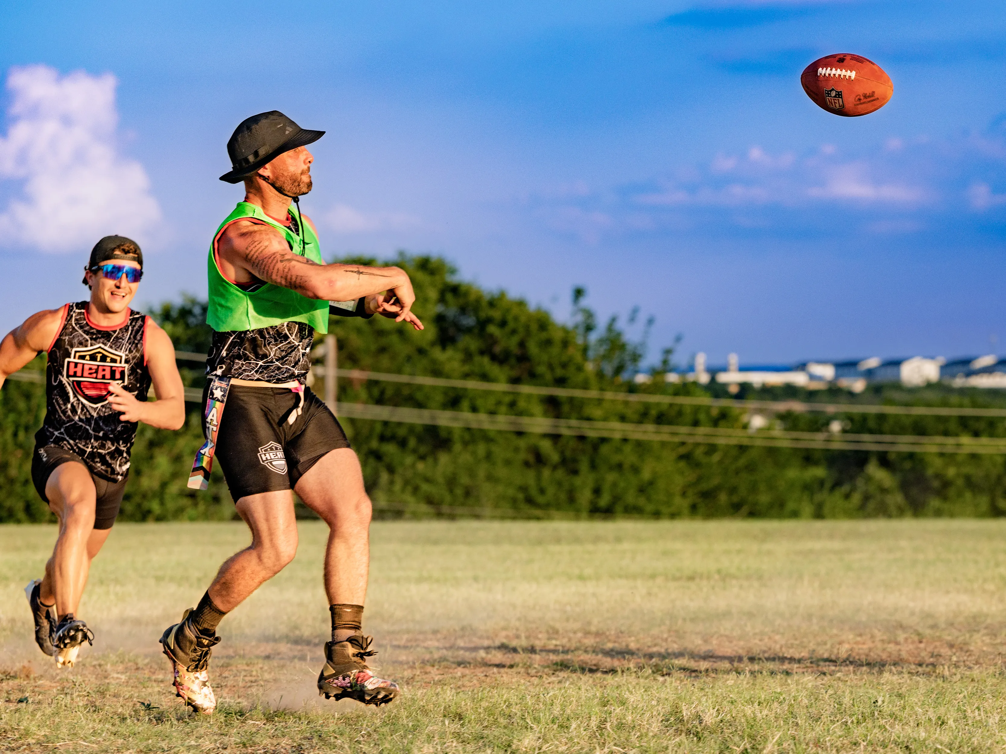 On a sunlit grassy field, an ATX Heat (Black) quarterback in a green scrimmage vest and bucket hat releases a pass while a teammate in a “HEAT” jersey and sunglasses sprints behind; both wear flag belts, with trees and a distant skyline under a bright blue sky during Gay Bowl 2025 practice.