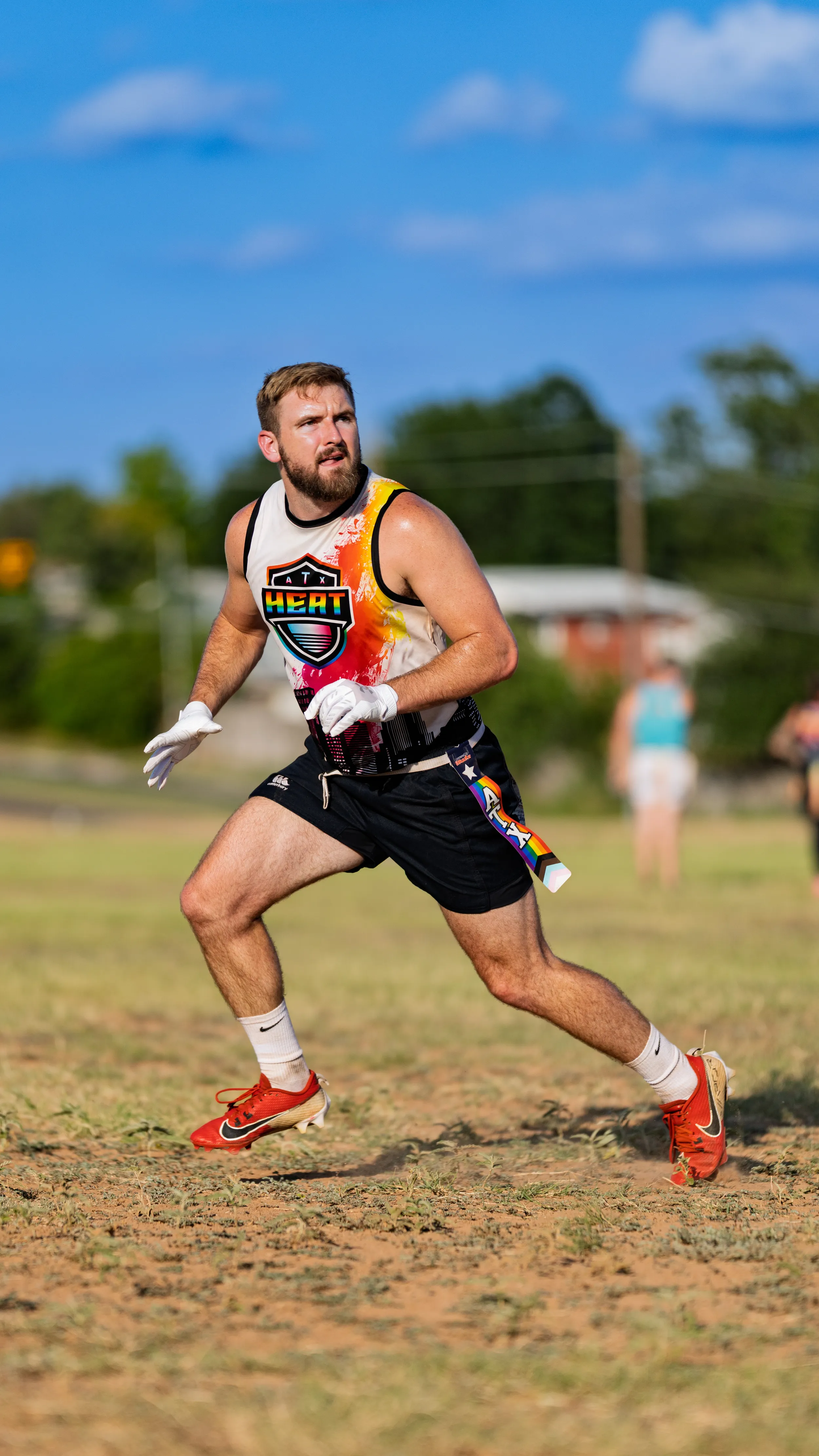 An ATX Heat (Black) flag football player runs on a sunny practice field, looking upfield, wearing a black jersey with a colorful HEAT logo, black shorts with a rainbow stripe, white gloves, red cleats, and a belt with pull flags. Blurred teammates and trees sit in the background during Gay Bowl 2025 practice.