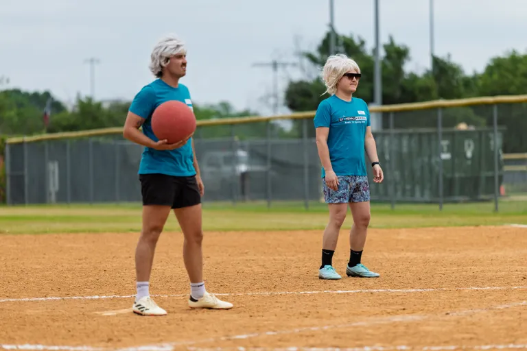 Two kickball players wearing light-colored wigs, blue team shirts, and shorts stand on a dirt field; one holds a red kickball, ready to play, while the other stands nearby, watching the game.