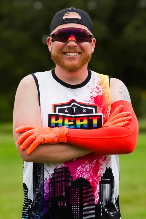 On a grassy practice field, an ATX Heat player stands smiling with arms crossed, wearing a white team jersey with a rainbow skyline and “ATX HEAT” logo, bright orange receiver gloves and matching arm sleeve, mirrored wraparound sunglasses, and a backward cap during Gay Bowl preparation.