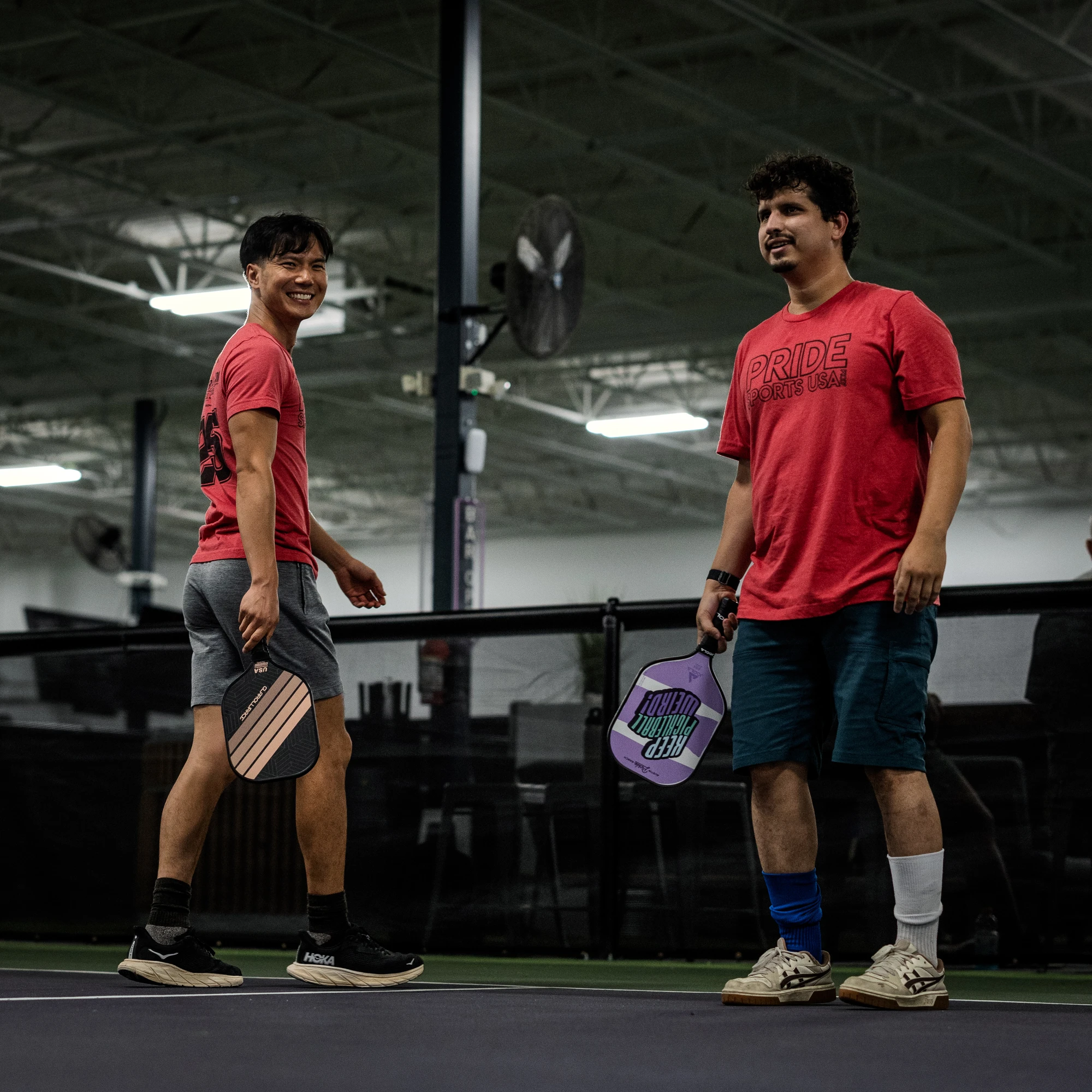 Two pickleball players stand on an indoor court, each holding a paddle and wearing athletic clothing, including red "PRIDE" shirts. They appear to be between points, with one player smiling toward the camera and a net visible behind them.