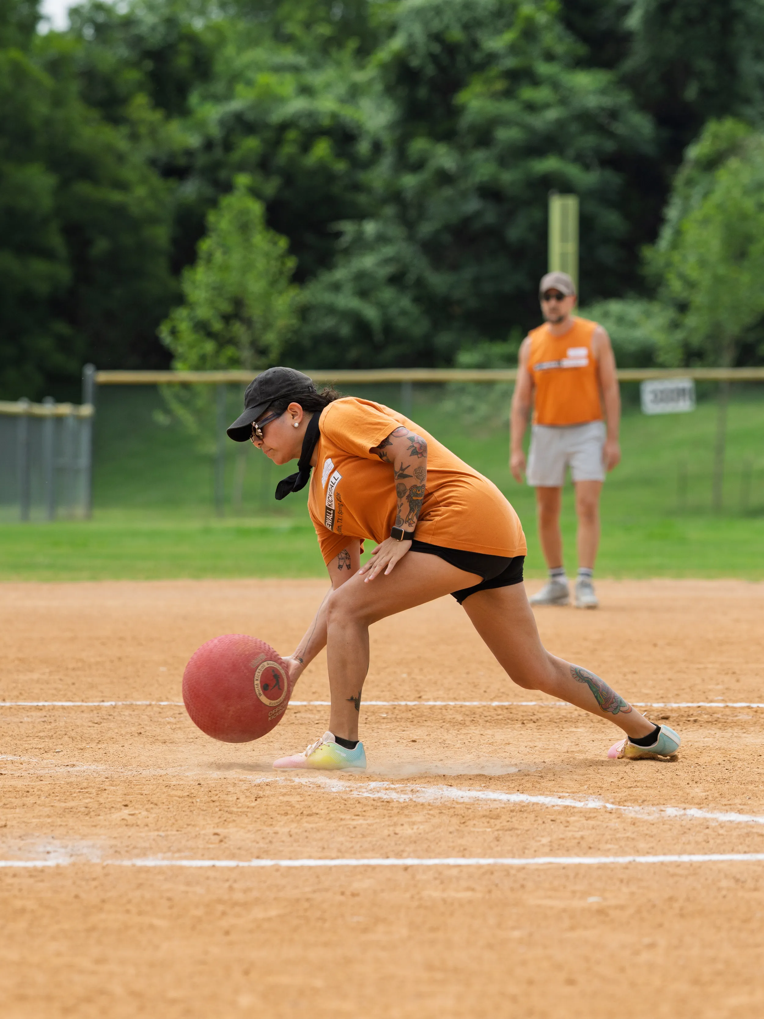 On a kickball field, a player in an orange team shirt, black cap, and sunglasses lunges low to scoop up a rolling red kickball along a chalked baseline. A teammate in a matching shirt watches from the infield, with fencing and trees in the green outfield behind.