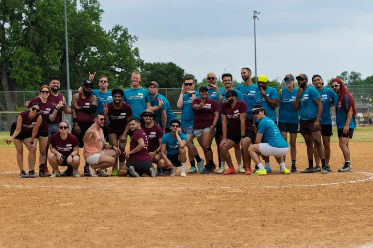 A group of kickball players wearing maroon and blue team shirts pose together on a dirt kickball field, some kneeling and others standing, with several making playful hand gestures or peace signs. Trees and part of the field’s fencing are visible in the background.