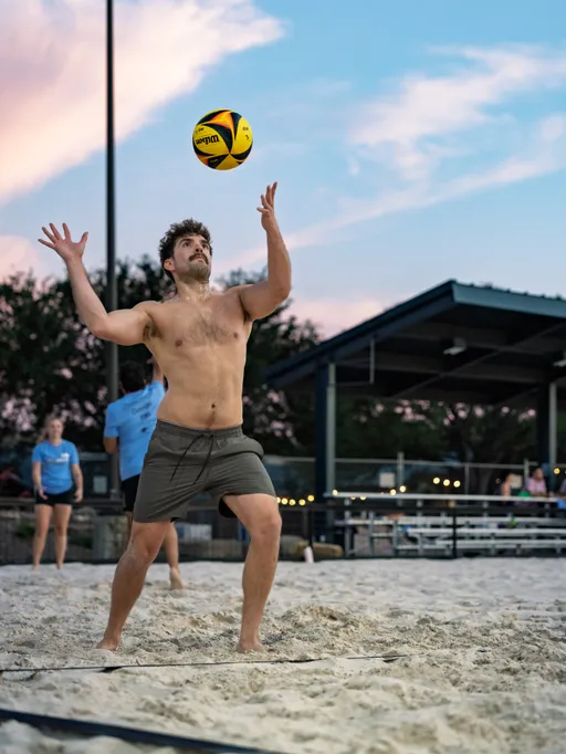 A volleyball player wearing shorts prepares to serve the ball on a sand court at sunset, with other players and a covered seating area visible in the background.