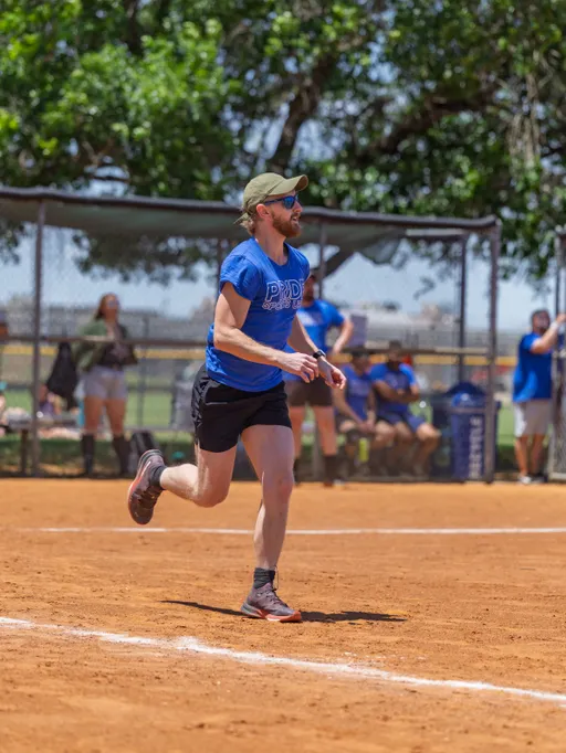 A player wearing a blue "PRIDE" team shirt, black shorts, a green cap, and athletic shoes is running on a dirt kickball field, with a fence, spectators, and trees visible in the background.