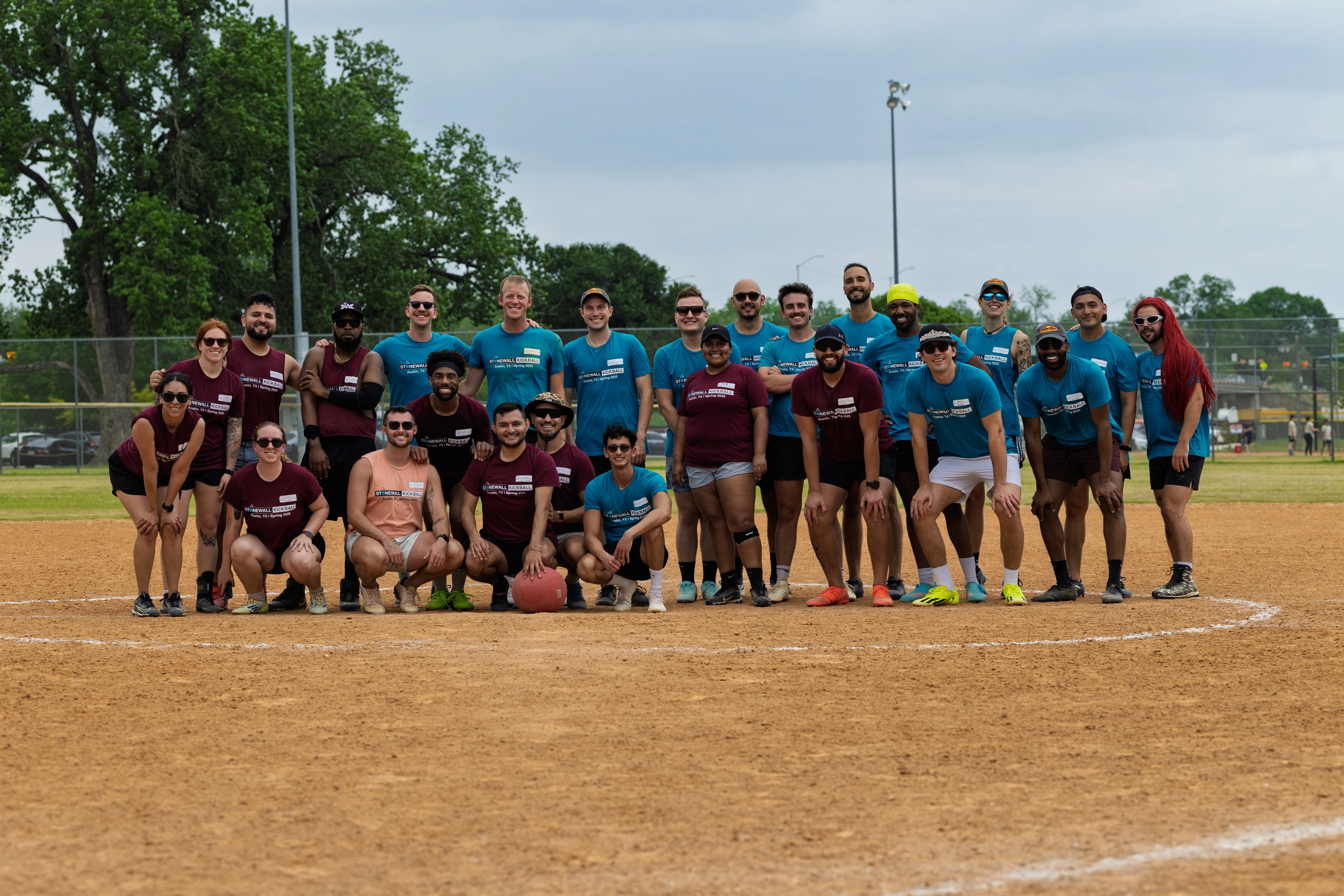 A group of kickball players wearing matching maroon and blue team shirts pose together on a kickball field, with one player holding a red kickball in front of them. The setting is outdoors on a dirt field with grass and trees visible in the background.