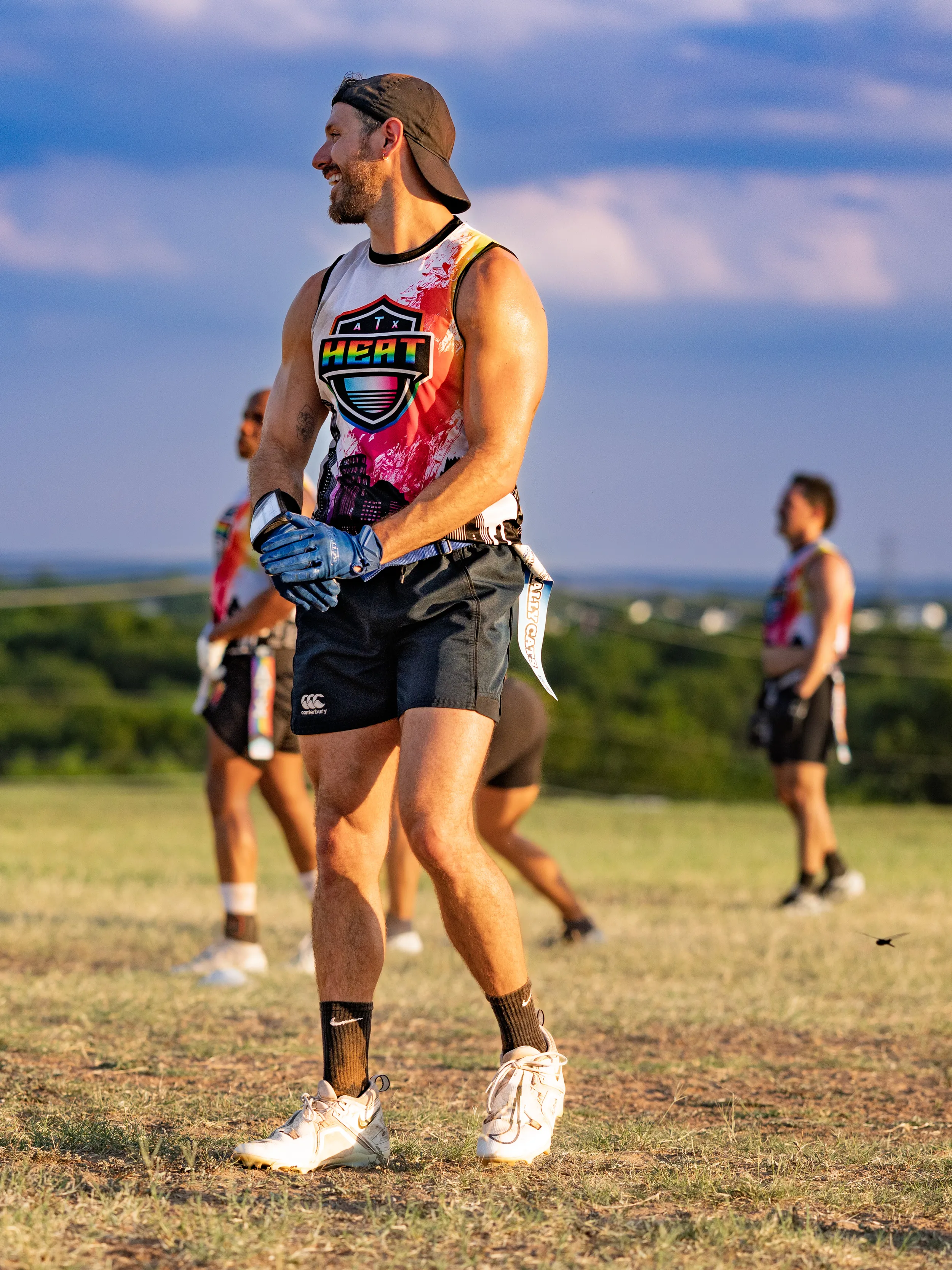ATX Heat (Black) players run a flag football practice for Gay Bowl 2025 on an open grassy field; a foreground player in a white-and-red HEAT jersey, black shorts, backward cap, blue gloves, cleats, and a flag belt pauses between plays while teammates in matching gear reset in the background under a bright, late-day sky.
