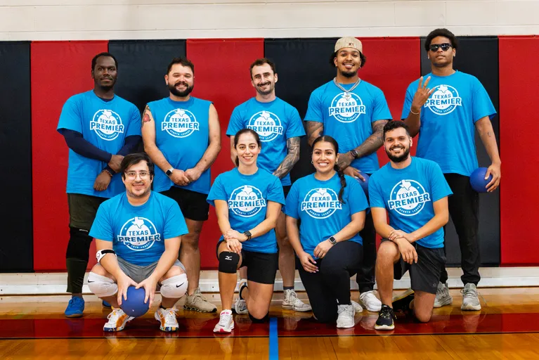 Team photo in a gym: nine dodgeball players in matching blue “Texas Premier” shirts kneel and stand on a polished court in front of red-and-black wall padding, some holding blue dodgeballs and wearing knee pads or athletic accessories, posed and ready for a game.