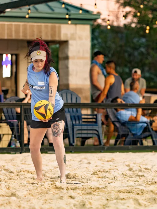 A volleyball player wearing a blue sleeveless shirt, black shorts, and a light-colored cap is serving a yellow and black volleyball on a sand court, with other athletes and spectators visible in the background.