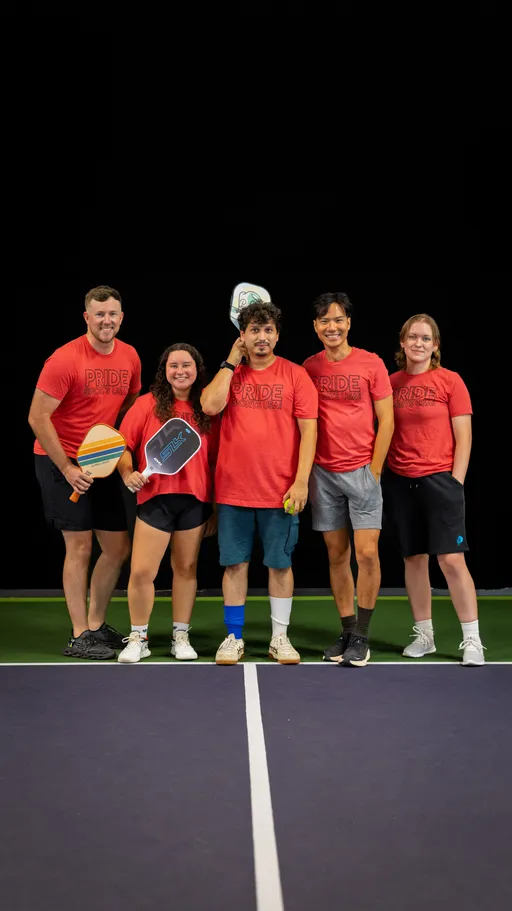 Five pickleball players stand together on an indoor court, each wearing matching red "Pride Pickleball" shirts and holding paddles, smiling and posing for a team photo.