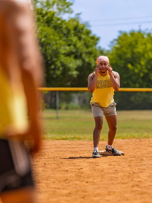 On a sunny kickball field, a player in a yellow “PRIDE” tank, gray shorts, bandana, cleats, and a white knee brace stands ready on the infield with hands cupped to call instructions. Another teammate in yellow is blurred in the foreground, with trees and a chain-link outfield fence behind.