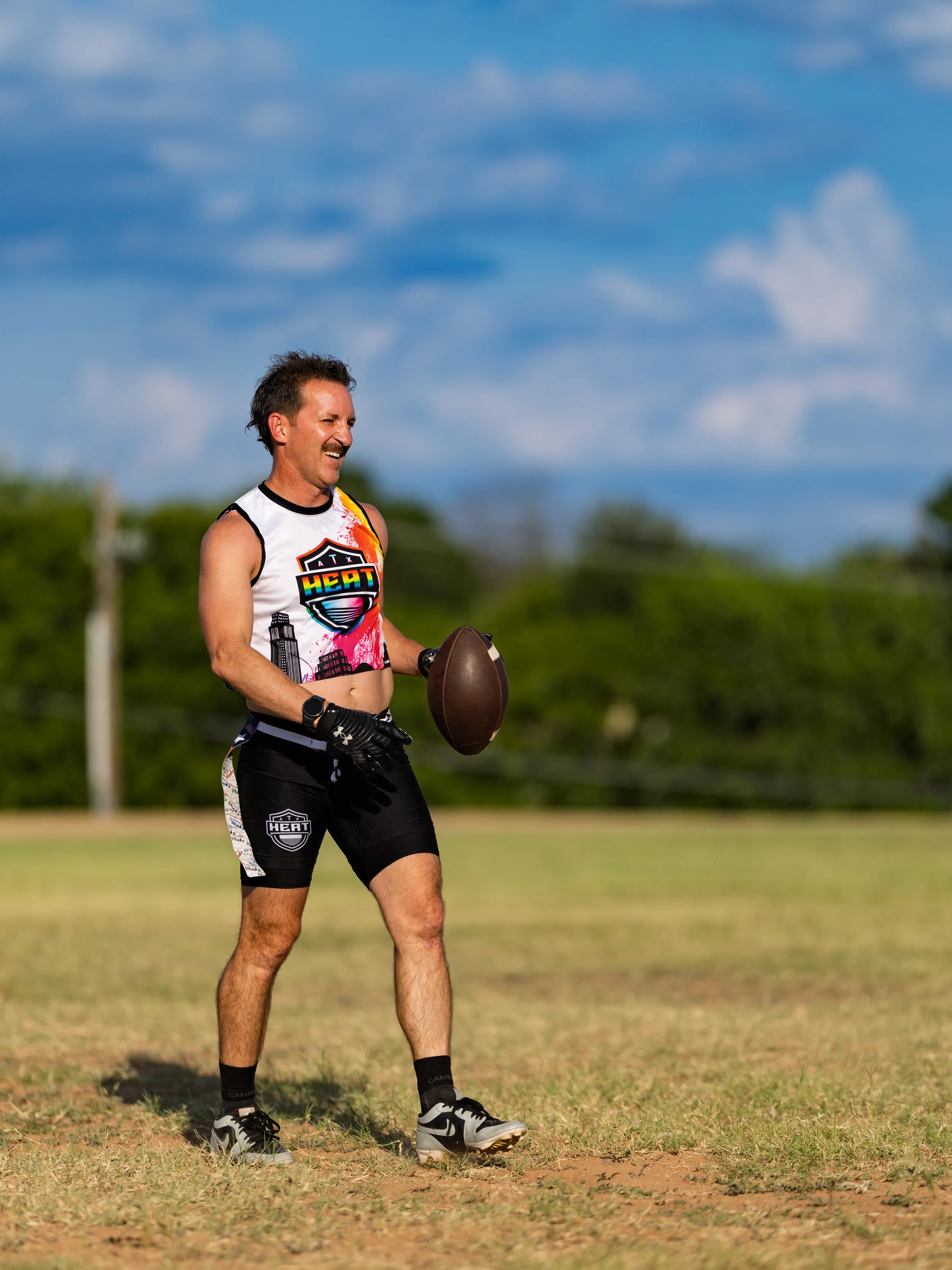 On a sunny grassy field, an ATX Heat (Black) player walks with a football in hand, smiling and wearing a sleeveless ATX Heat jersey with a colorful graphic, black compression shorts with team logos, black gloves, and cleats. Trees and blue sky frame the practice scene for Gay Bowl 2025.