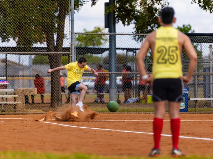 A player in a yellow “Pitch Please” shirt slides into a base along the baseline, kicking up a cloud of dirt as a green kickball rolls nearby. In the foreground, a teammate wearing a yellow jersey with the number 25 and red knee-high socks watches on a fenced kickball field.