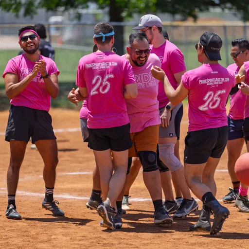 Players wearing matching bright pink shirts and athletic shorts celebrate together on the dirt of a sunny kickball field, with some clapping and others smiling as they gather in a group.