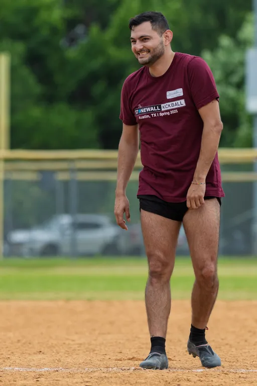 A player wearing a maroon "Stonewall Kickball" t-shirt, black athletic shorts, and sneakers stands on a kickball field, appearing relaxed and smiling, with grassy areas and a fence in the background.