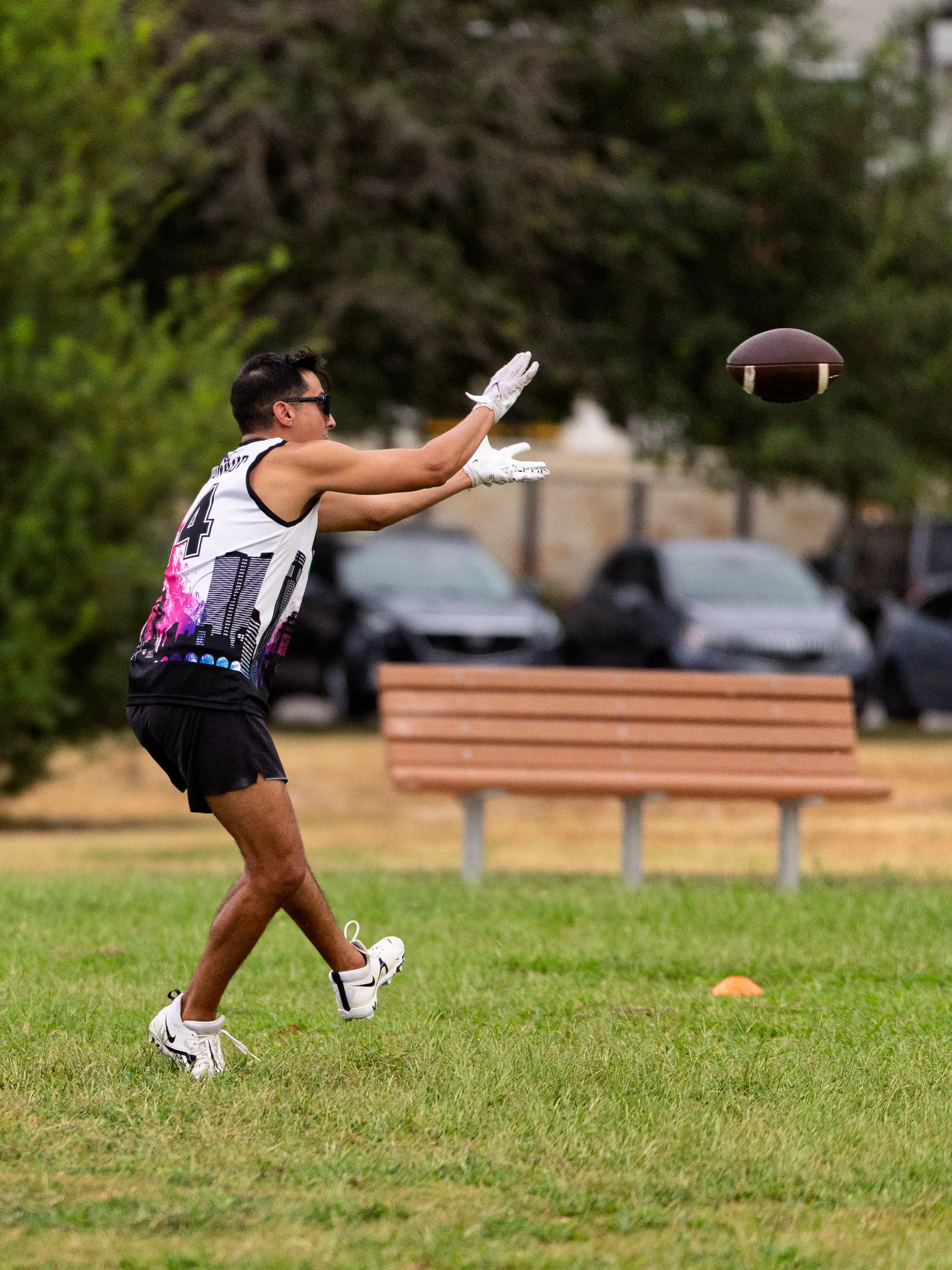 An ATX Heat (Orange) player in a sleeveless white-and-black jersey with the number 4, black shorts, white cleats, gloves, and sunglasses reaches forward to catch a football during practice drills on a grassy park field. An orange cone marks the area, with a bench and parked cars in the background.