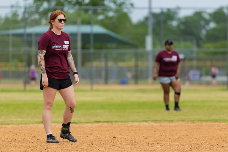 Two kickball players stand on a dirt field, both wearing matching maroon Stonewall Kickball shirts and athletic shorts, appearing focused as they watch the action during a game. The setting is an outdoor kickball field with grass, chain-link fences, and other players visible in the background.