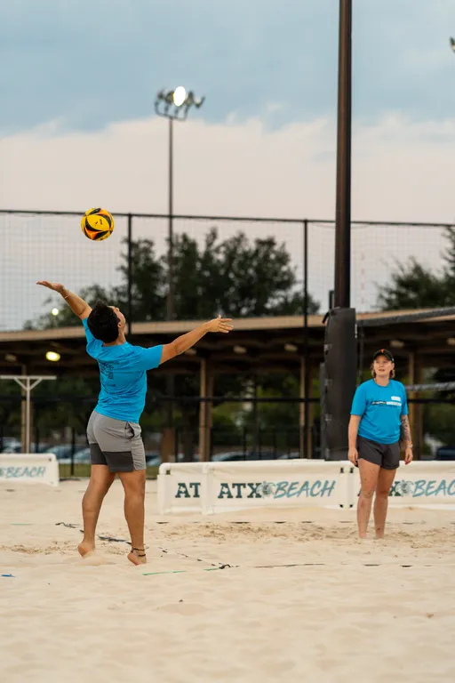 At dusk on an outdoor sand volleyball court, a player in a blue T-shirt and gray shorts tosses a yellow-and-black ball for a serve while a teammate in similar gear, wearing a cap, stands ready. The ATX Beach net, fencing, and field lights frame the sandy setting.