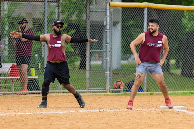 Two kickball players wearing matching maroon Stonewall Kickball shirts are on a dirt field near a base; one player is mid-stride with arms extended, while the other stands with hands on hips and a tattoo on one leg. A chain-link fence, additional players, and sports equipment can be seen in the background.