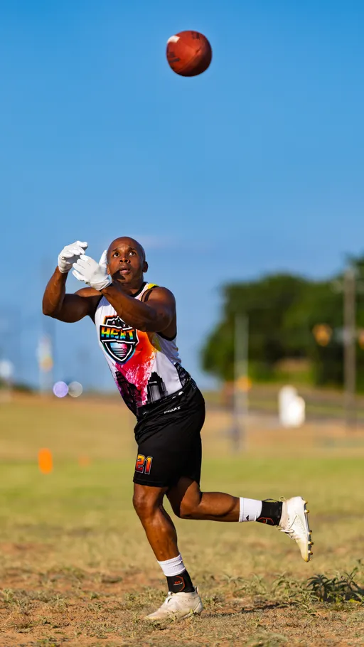 On a sunny flag football practice field, an ATX Heat (Black) player in a colorful “HEAT” jersey, black shorts with the number 21, white gloves, and cleats reaches up to secure a pass as the football descends. Orange cones dot the open grassy area in the background, indicating team drills for Gay Bowl 2025.