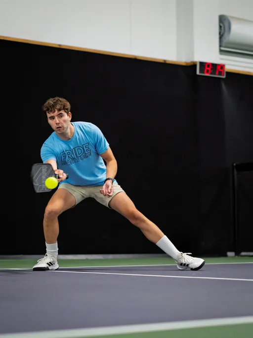A pickleball player wearing a blue "Pride Pickleball" T-shirt, light shorts, and athletic shoes stretches to hit a ball with a paddle on an indoor court.