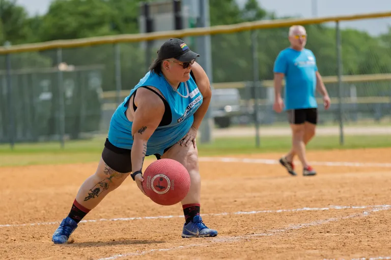 A kickball player in a blue team shirt and cap is crouched down, preparing to roll the ball, while another player stands in the background on a sandy kickball field.