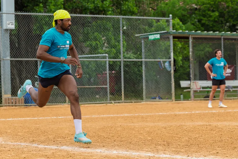 A player wearing a bright yellow bandana, turquoise Stonewall Kickball shirt, black shorts, and turquoise shoes is running across a dirt kickball field, while another player in similar attire stands near the fence in the background.