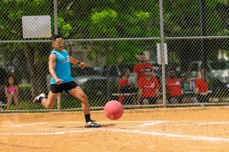 A kickball player in a blue athletic tank top and sunglasses is captured mid-kick on a sandy kickball field, with spectators in orange shirts watching from behind a chain-link fence.