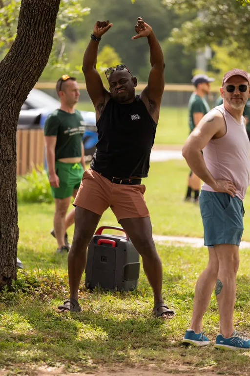 A player in a black tank top and pink shorts stands on grass with arms raised and expressive body language, while another person in sunglasses and a cap stands nearby with hands on hips; other players and a kickball field are visible in the background.