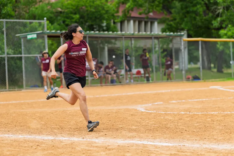 A player wearing a maroon Stonewall Kickball shirt, black shorts, and athletic shoes is running across a dirt kickball field, with teammates and spectators visible in the fenced dugout area in the background.