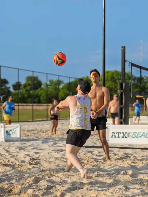 Two sand volleyball players are focused on the ball, with one preparing to bump it while wearing a tie-dye tank top, shorts, and a cap; the game takes place on a sunny sand court surrounded by a fence, with other players and a net visible in the background.
