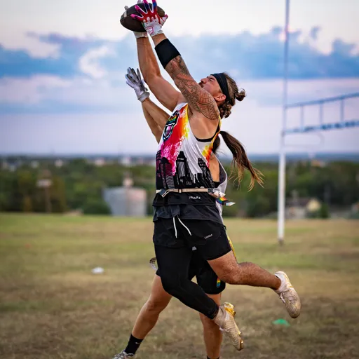 During ATX Heat (Black) practice for Gay Bowl 2025, a player leaps on an open field to grab a football with both hands, wearing a white tank with pink-and-black graphics, black shorts, gloves, arm bands, and cleats; a goal frame and distant trees sit beneath a cloudy sunset.