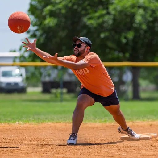A player on a kickball field, wearing an orange team shirt, dark shorts, a cap, and sunglasses, is reaching out with both hands to catch a kickball in mid-air.