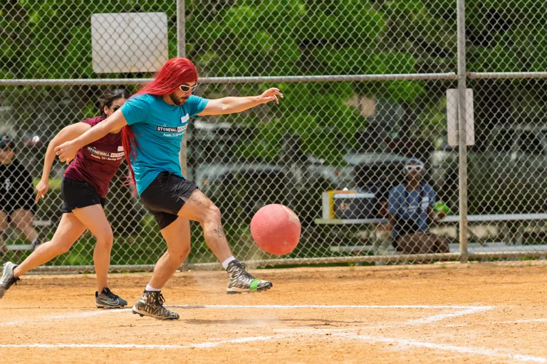 A player wearing a vibrant wig, sunglasses, a blue Stonewall Kickball shirt, and black shorts is kicking a red kickball on a dirt field while another competitor in a maroon shirt and black shorts runs closely behind; both are on a fenced kickball field with green trees and blurred spectators in the background.