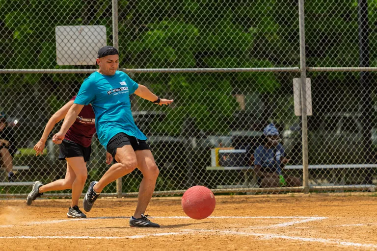 A player in a bright blue shirt and black headband is kicking a red kickball on a sandy kickball field, while another player in a maroon shirt runs behind them. A chain-link fence and green trees are visible in the background.