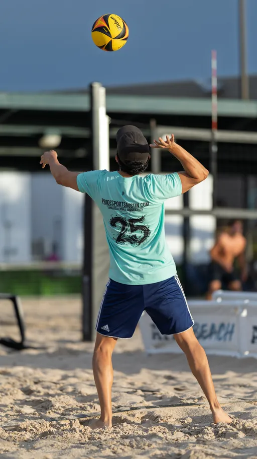 A volleyball player in a light blue shirt, navy shorts, and a black cap is preparing to serve the ball on a sand volleyball court, with the ball airborne above their hand and other players visible in the background.