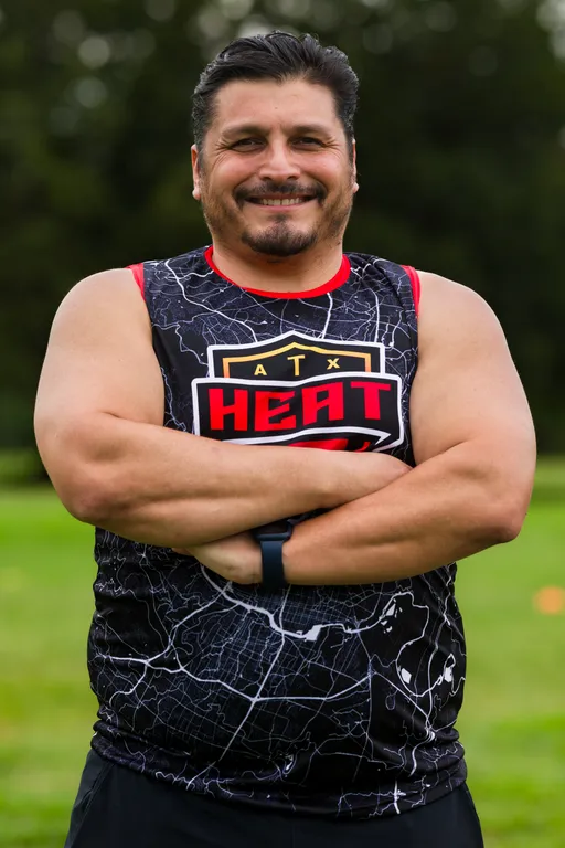An ATX Heat player stands on a grassy flag football practice field with trees in the background, arms crossed and smiling, wearing a black sleeveless ATX HEAT jersey with red accents and a map-like pattern, plus a fitness watch.