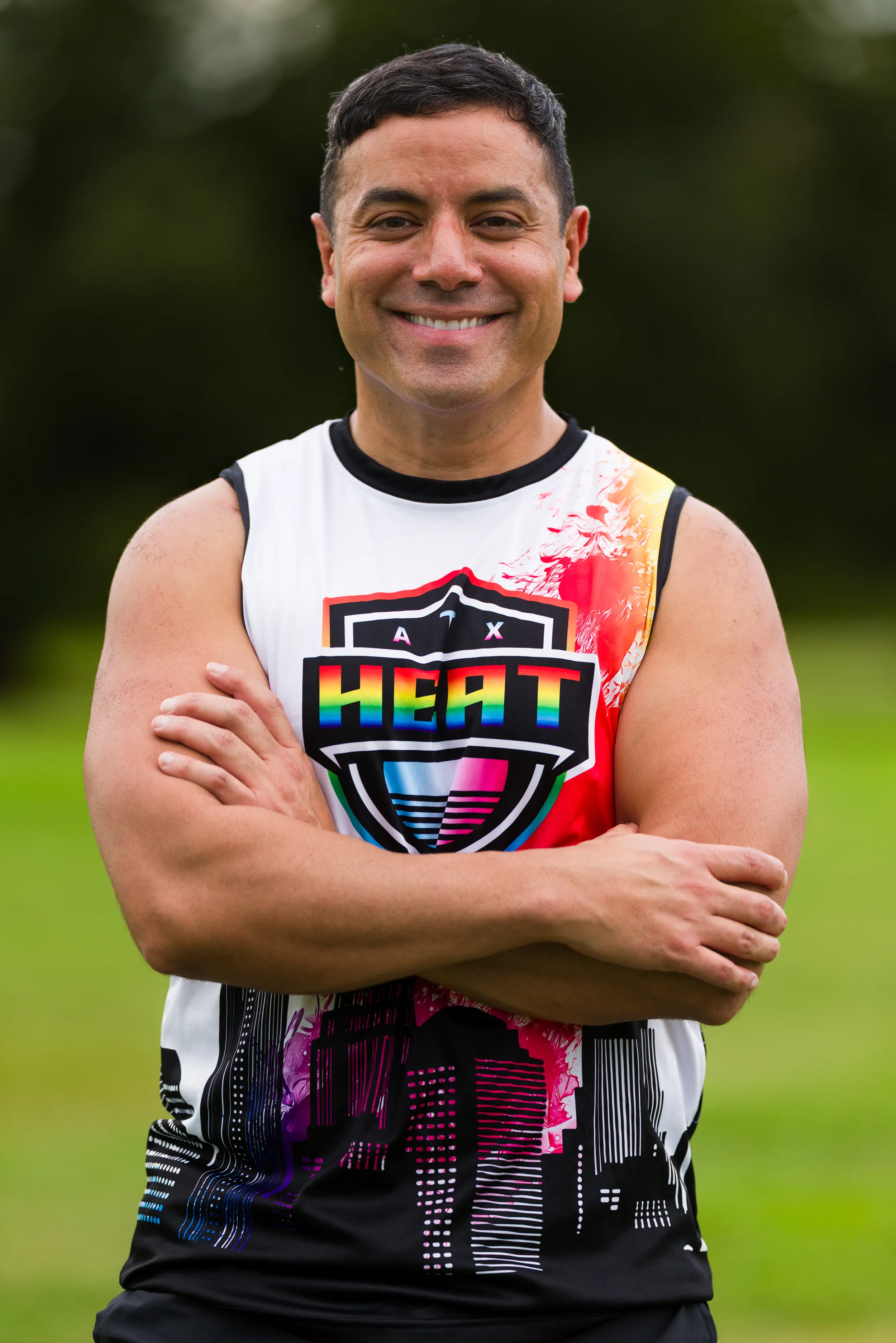 An ATX Heat player stands on a grassy practice field with arms crossed, wearing a sleeveless team jersey featuring a rainbow “HEAT” shield and abstract cityscape pattern, with blurred trees in the background during Gay Bowl 2025 preparations.