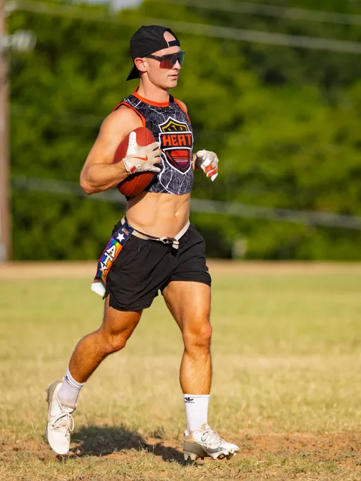 ATX Heat (Black) player runs across a sunny grass field during practice for Gay Bowl 2025, cradling a football and looking ahead. The athlete wears a cropped “HEAT” jersey, black shorts with a flag belt, white cleats, receiver gloves, a backward cap, and sunglasses.
